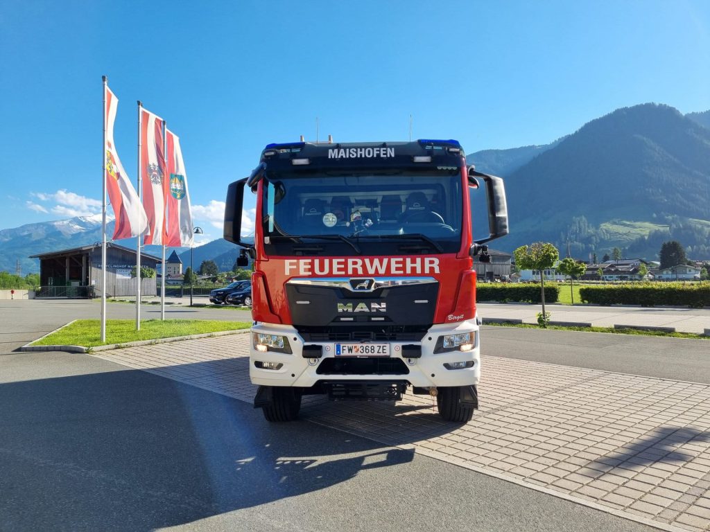 Feuerwehrfahrzeug der FF Maishofen von vorn vor Fahnen und Bergkulisse bei blauem Himmel