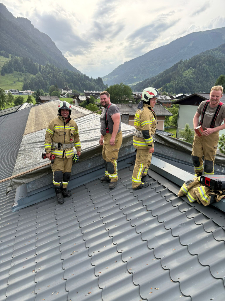 Feuerwehrleute stehen auf einem Metalldach neben einer provisorisch abgedeckten Dachfläche im Gebirgstal