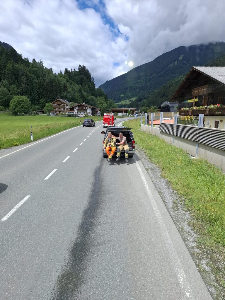 Zwei Feuerwehrleute sitzen an einer Straße neben einer dunklen Flüssigkeitsspur, ein Feuerwehrfahrzeug steht voraus